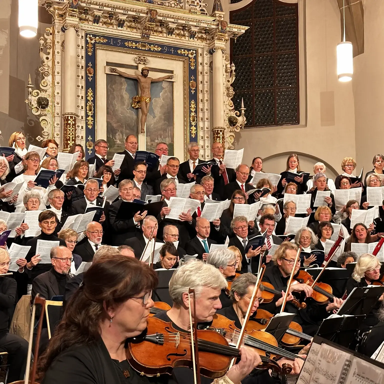Ein Orchester im Vordergrund in einer Kirche. Im Hintergrund festlich gekleideten Sängerinnen und Sängern im Chor. 