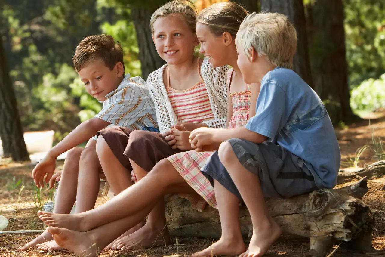 Vier Kinder im Alter etwa von 10 Jahren sitzen auf einem umgesägten Baum im Wald. Es ist Sommer.