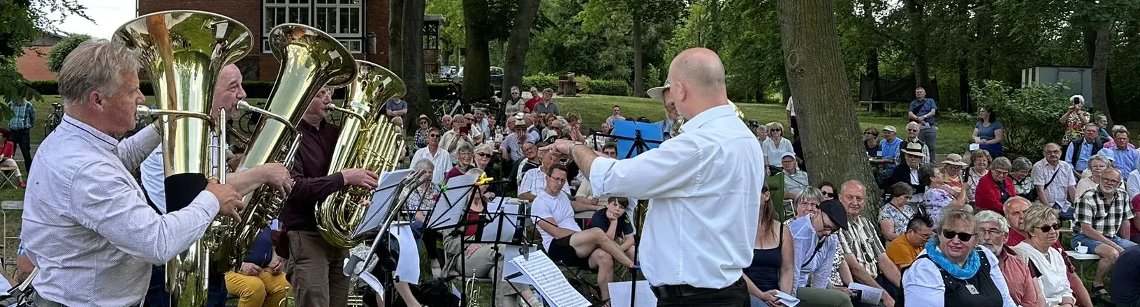 Im Vordergrund drei Männer mit einer Tuba, die von einem Dirigenten angeleitet werden. Im Hintergrund eine Gottesdienstgesellschaft im Freien in einer parkähnlichen Landschaft.