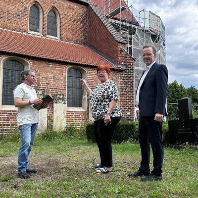 Drei Menschen stehen vor einem Kirchengebäude, das in Teilen eingerüstet ist. Das Gebäude ist aus Backstein.
