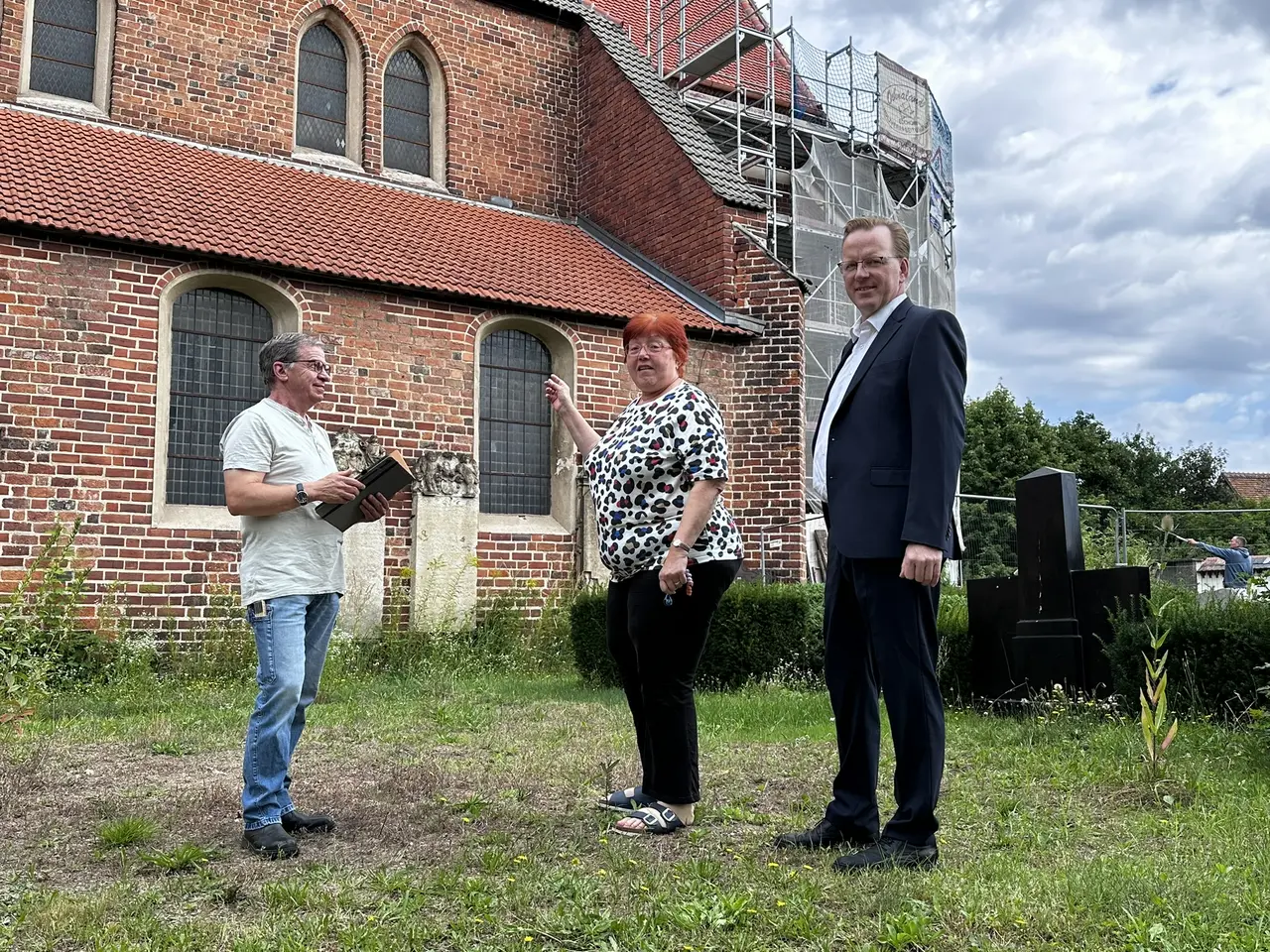 Drei Menschen stehen vor einem Kirchengebäude, das in Teilen eingerüstet ist. Das Gebäude ist aus Backstein.