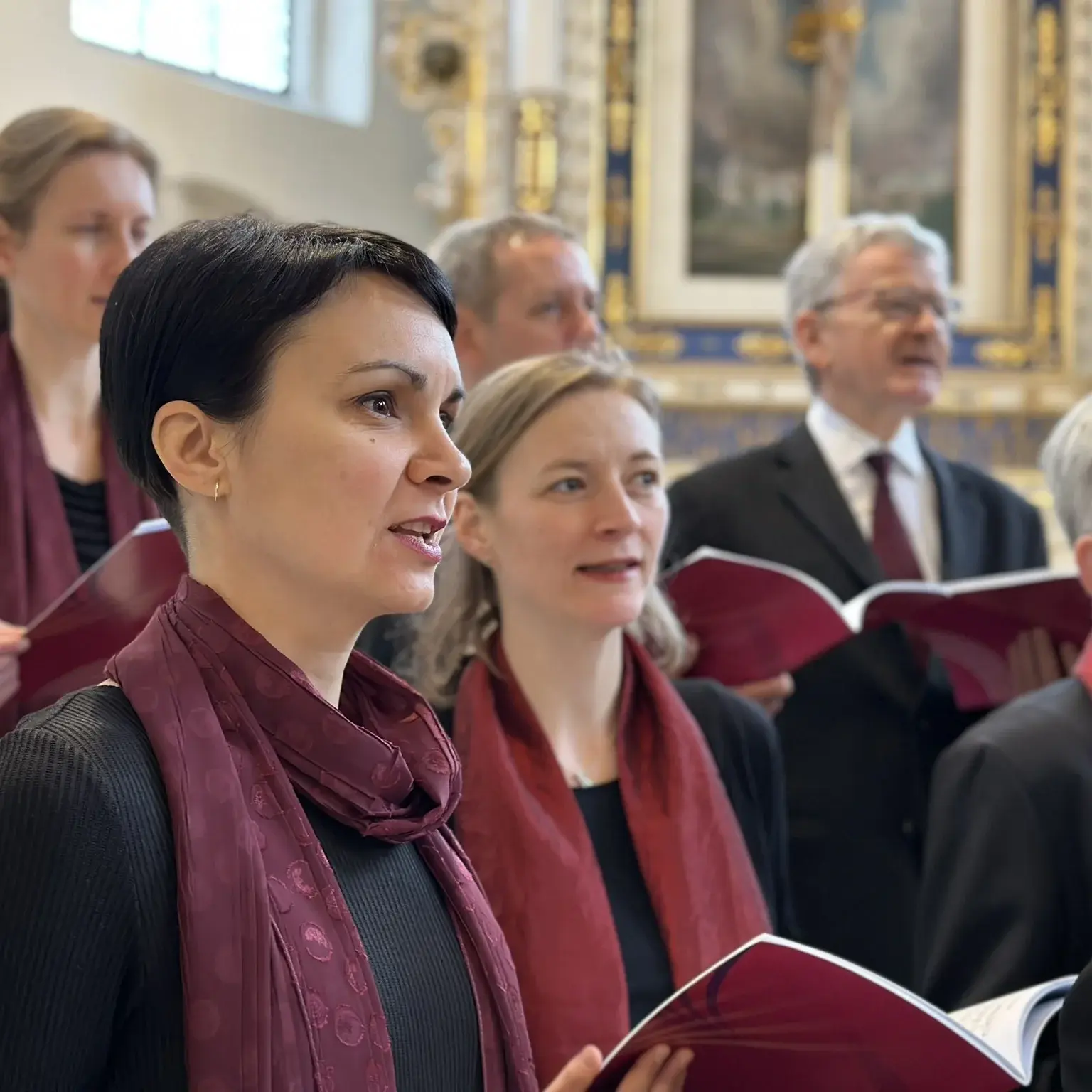 Drei Frauen in einheitlicher Kleidung in schwarz mit einem weinroten Schal stehen vor einem Altar. Sie singen.