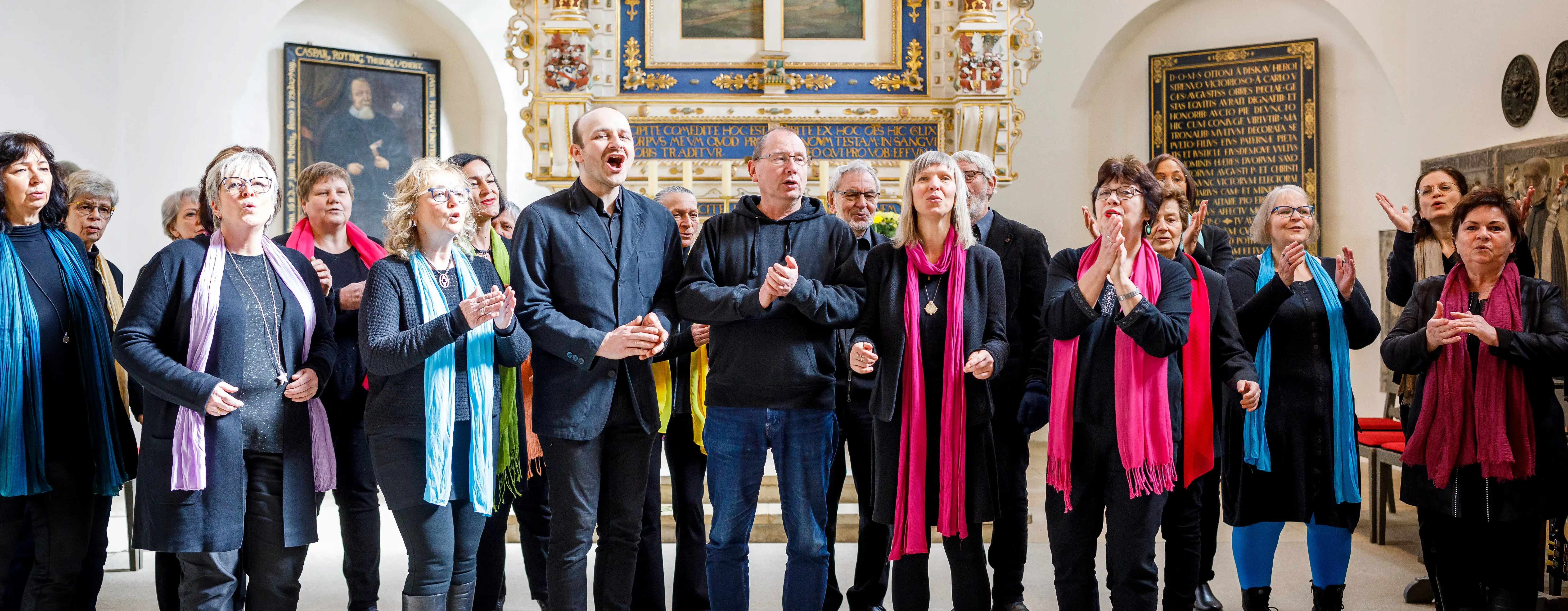 Ein Chor vor einem Altar in einer Kirche. Die Menschen singen. Sie tragen bunte Schals. 